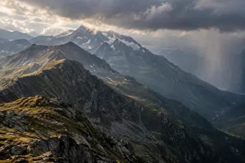 Rocky mountain ridges with warm patches of grass beneath dark clouds and a distant rain curtain.