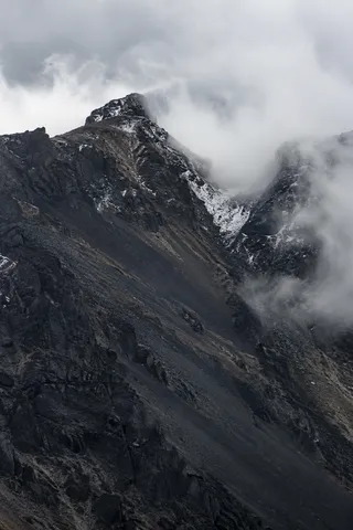 Portrait view of a dark craggy ridge and snow-streaked saddle partly hidden by white cloud.