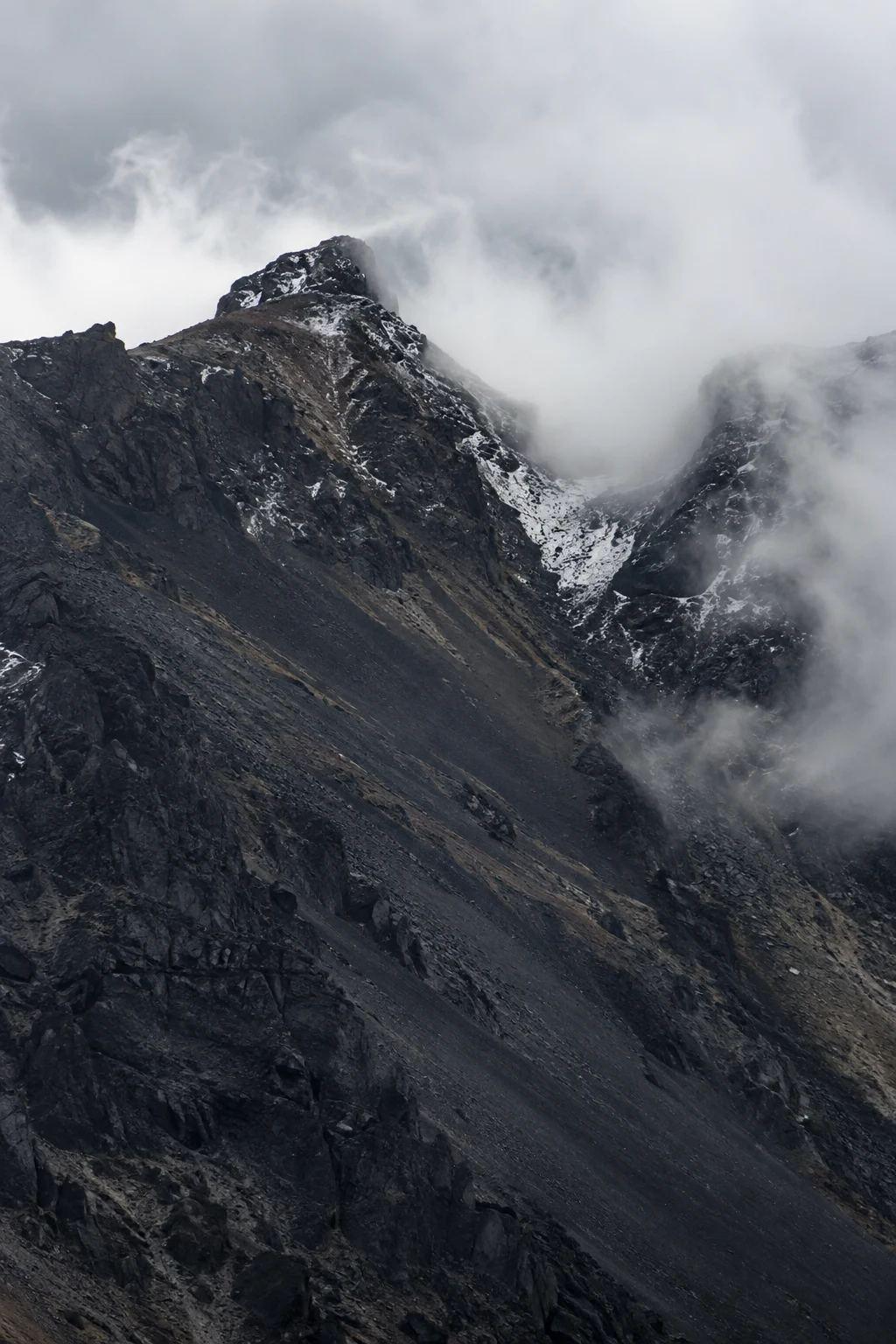 Portrait view of a dark craggy ridge and snow-streaked saddle partly hidden by white cloud.