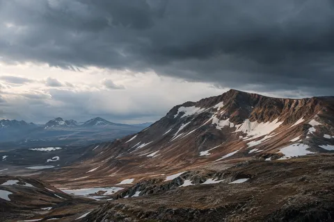 Broad reddish mountain ridge with irregular snow patches under a dark cloud shelf.
