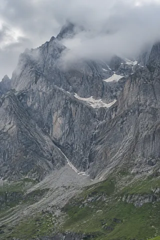 Tall gray granite mountain face with sheltered snow ledges and a green rocky apron under cloud.