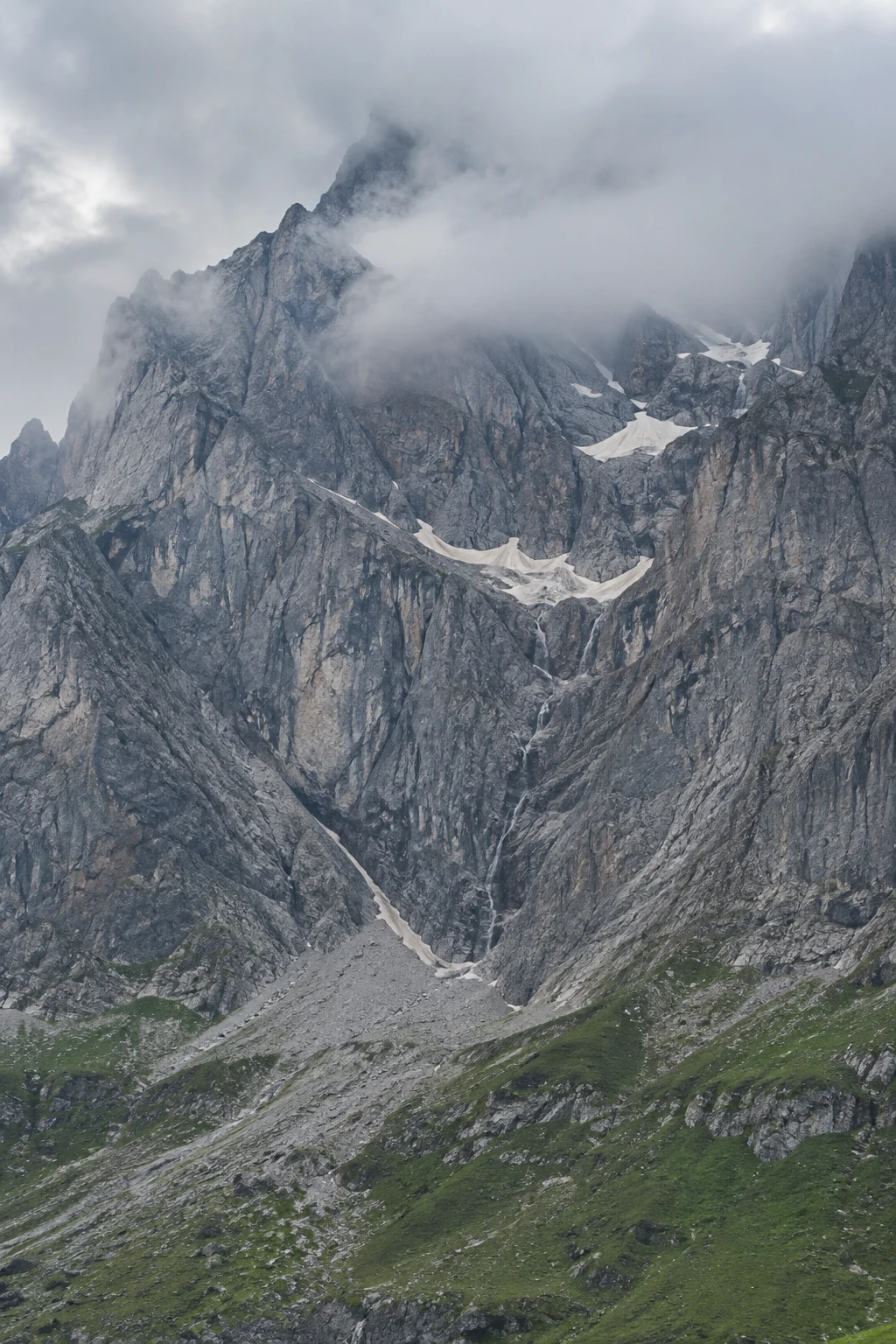 Tall gray granite mountain face with sheltered snow ledges and a green rocky apron under cloud.