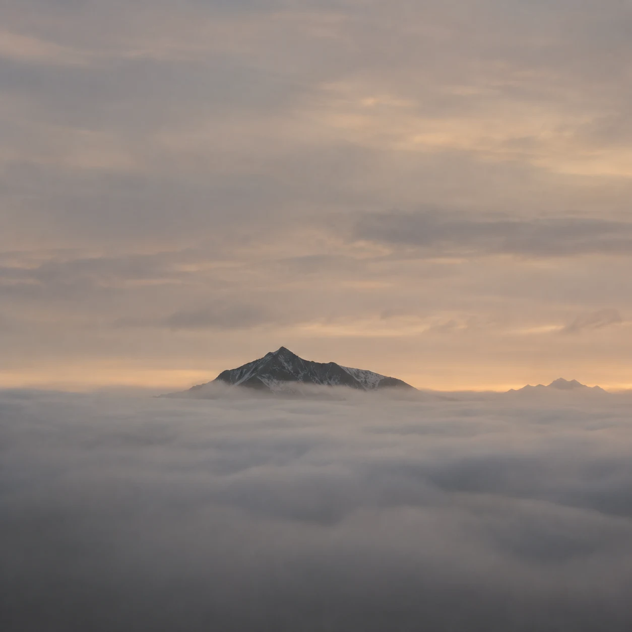 Small dark mountain peak above a broad fog bank under a warm gray evening sky.