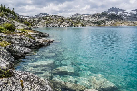 Turquoise alpine lake with submerged rocks, an irregular granite shoreline and low snowy hills.