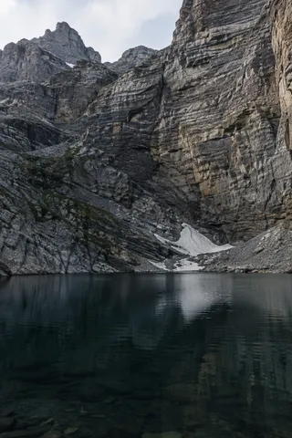 Dark mountain pool reflecting a layered cliff and a sheltered patch of snow.