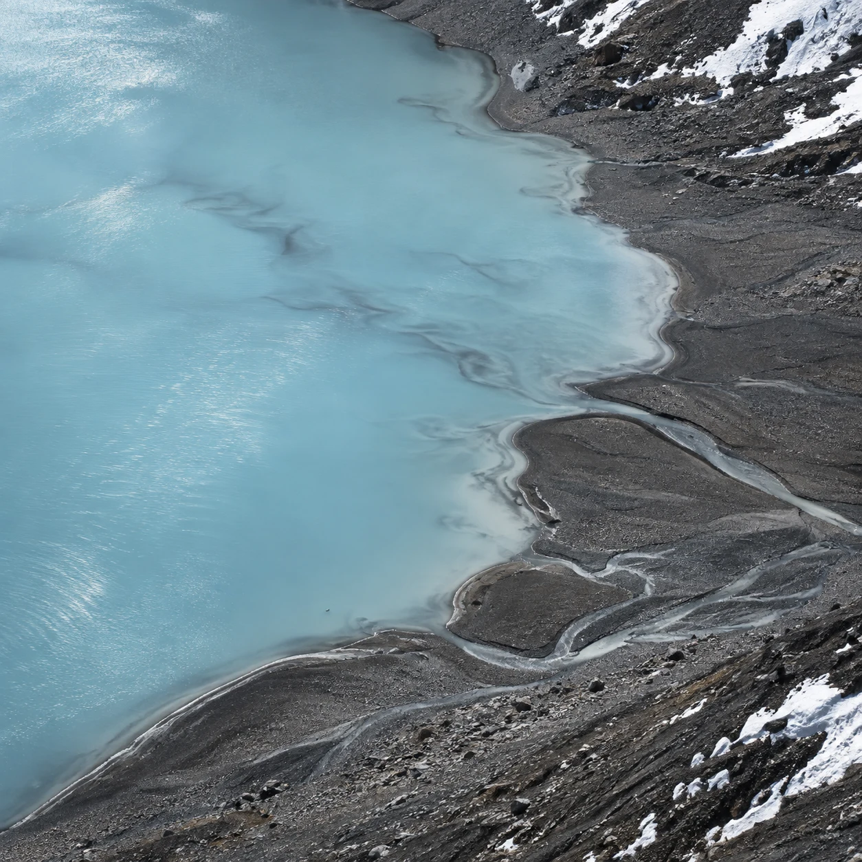 Elevated view of milky blue water curving against dark gravel and small branching channels.