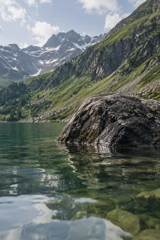 Large wet gray boulder partly submerged in a clear mountain lake with green slopes behind.