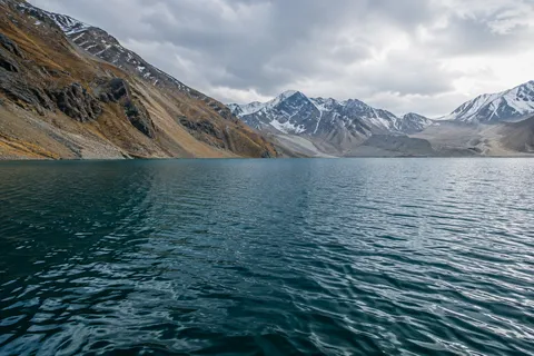 Wide teal alpine lake with fine ripples, tawny slopes at left and pale snowy mountains beyond.