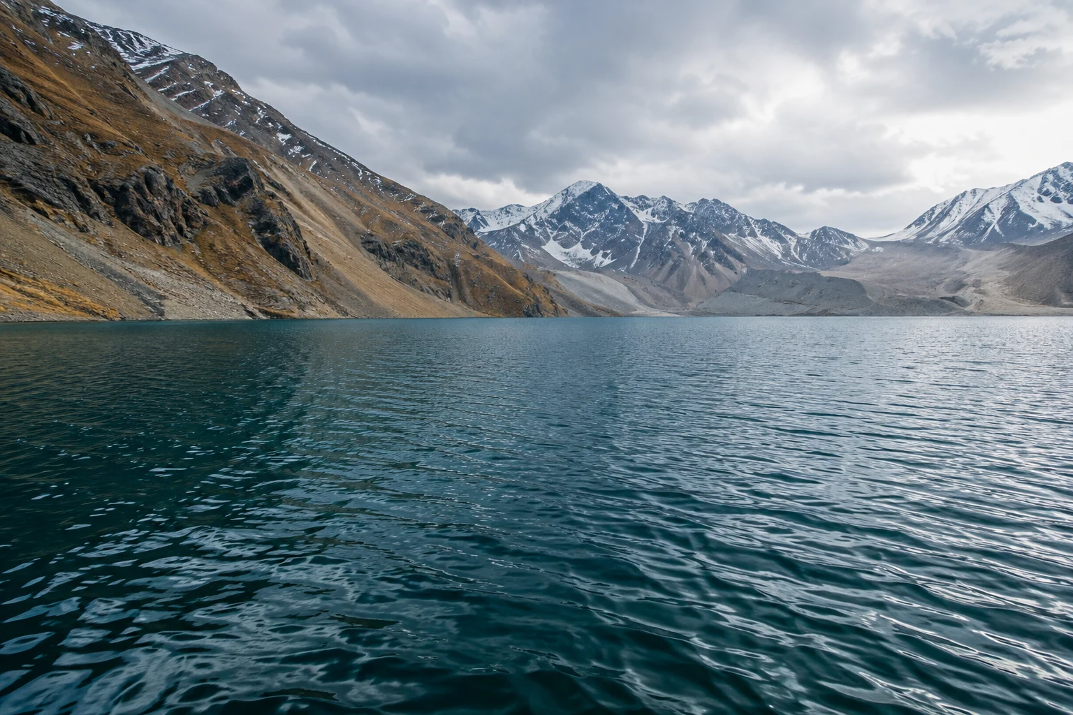 Wide teal alpine lake with fine ripples, tawny slopes at left and pale snowy mountains beyond.