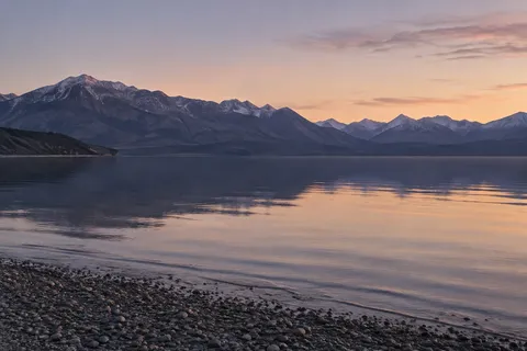 Soft peach dawn sky above a violet mountain ridge reflected in a calm lake beside a pebble beach.