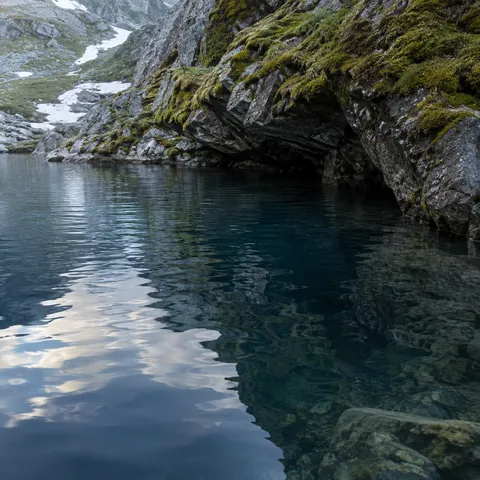 Quiet blue alpine cove beneath rough mossy rock with rippled reflections and visible underwater stones.