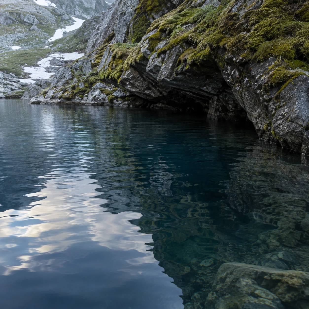 Quiet blue alpine cove beneath rough mossy rock with rippled reflections and visible underwater stones.