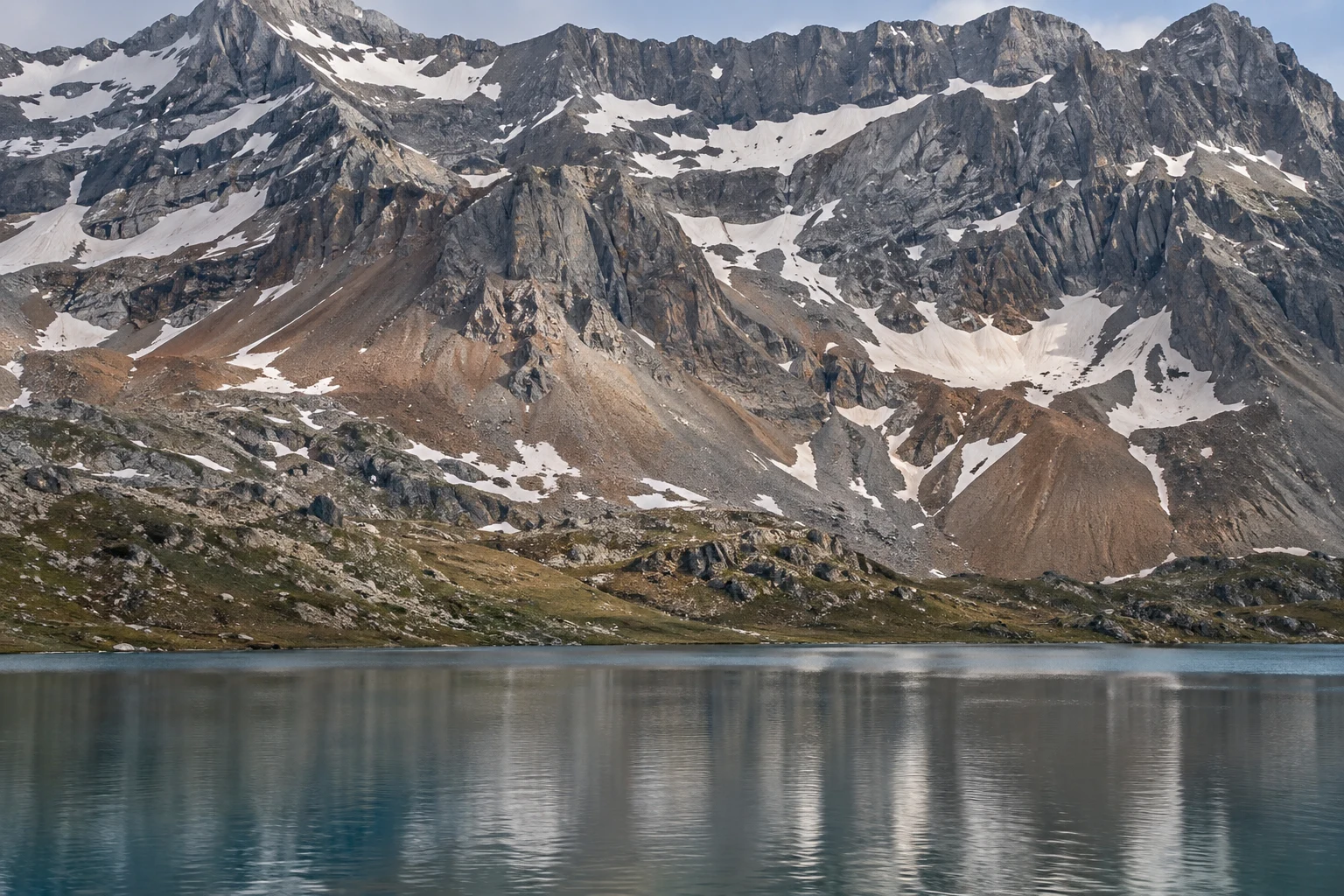 Wide gray mountain wall with melting snow patches, brown scree fans and a softly reflecting lake.