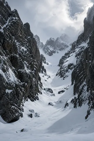 Narrow snow-filled mountain couloir between two steep dark rocky walls.