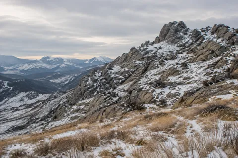 Low rocky mountain ridge lightly covered in snow above golden dry grasses and distant blue hills.