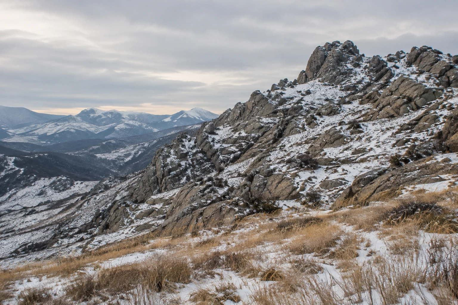 Low rocky mountain ridge lightly covered in snow above golden dry grasses and distant blue hills.
