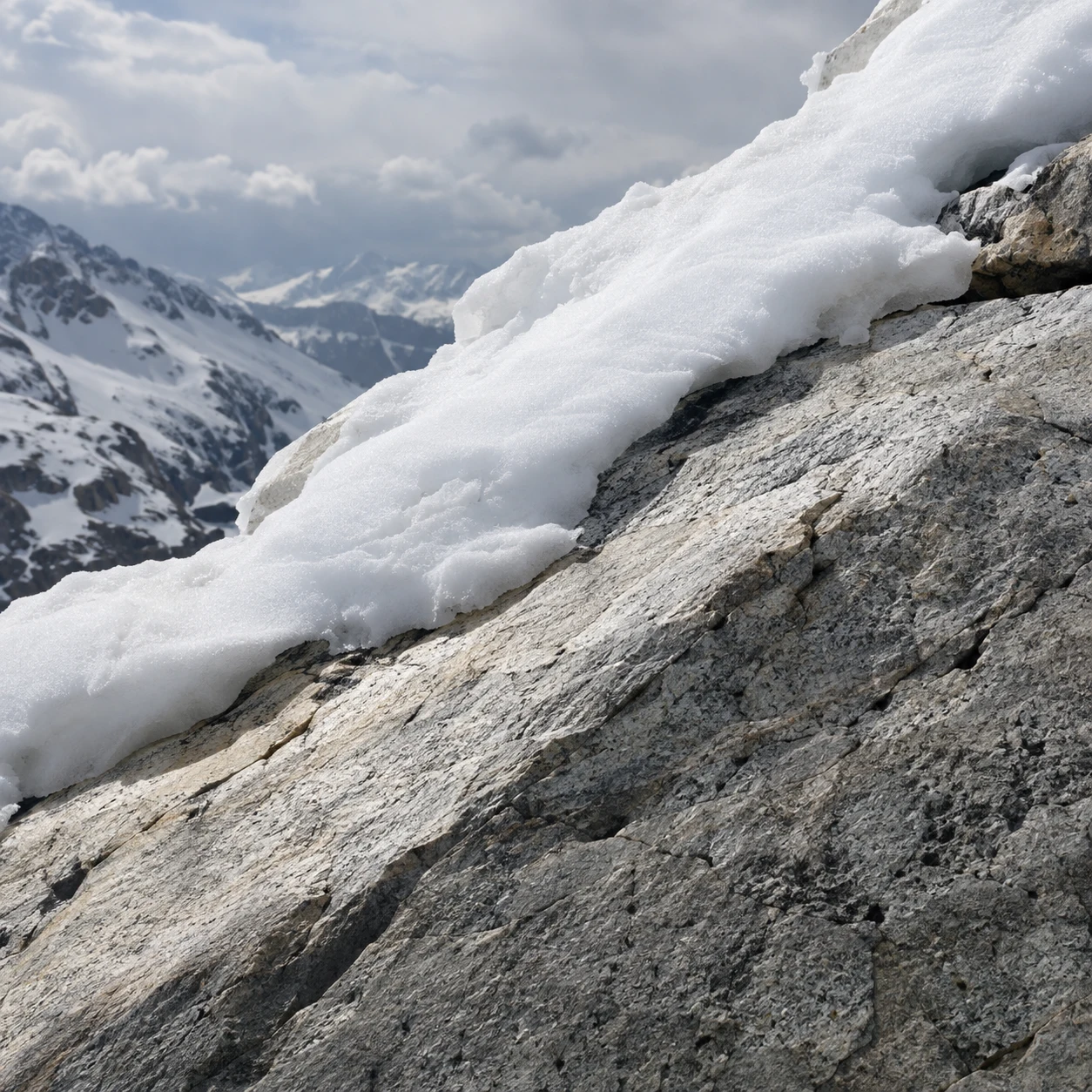 Close diagonal pale granite slab topped by granular snow, with soft distant snowy mountains.