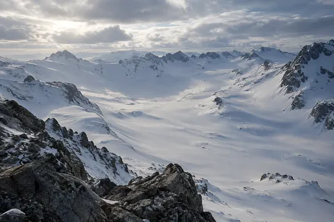 Wide empty snow basin surrounded by low rugged peaks, viewed over dark foreground rocks.