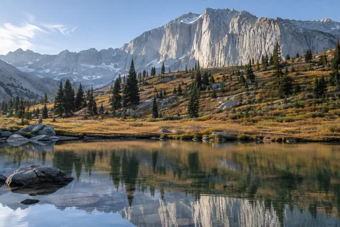 Dark conifers on a golden mountain slope beneath pale granite, reflected in a foreground pool.