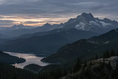 Dark conifer-covered mountain layers surrounding a pale lake beneath a rugged ridge at dusk.