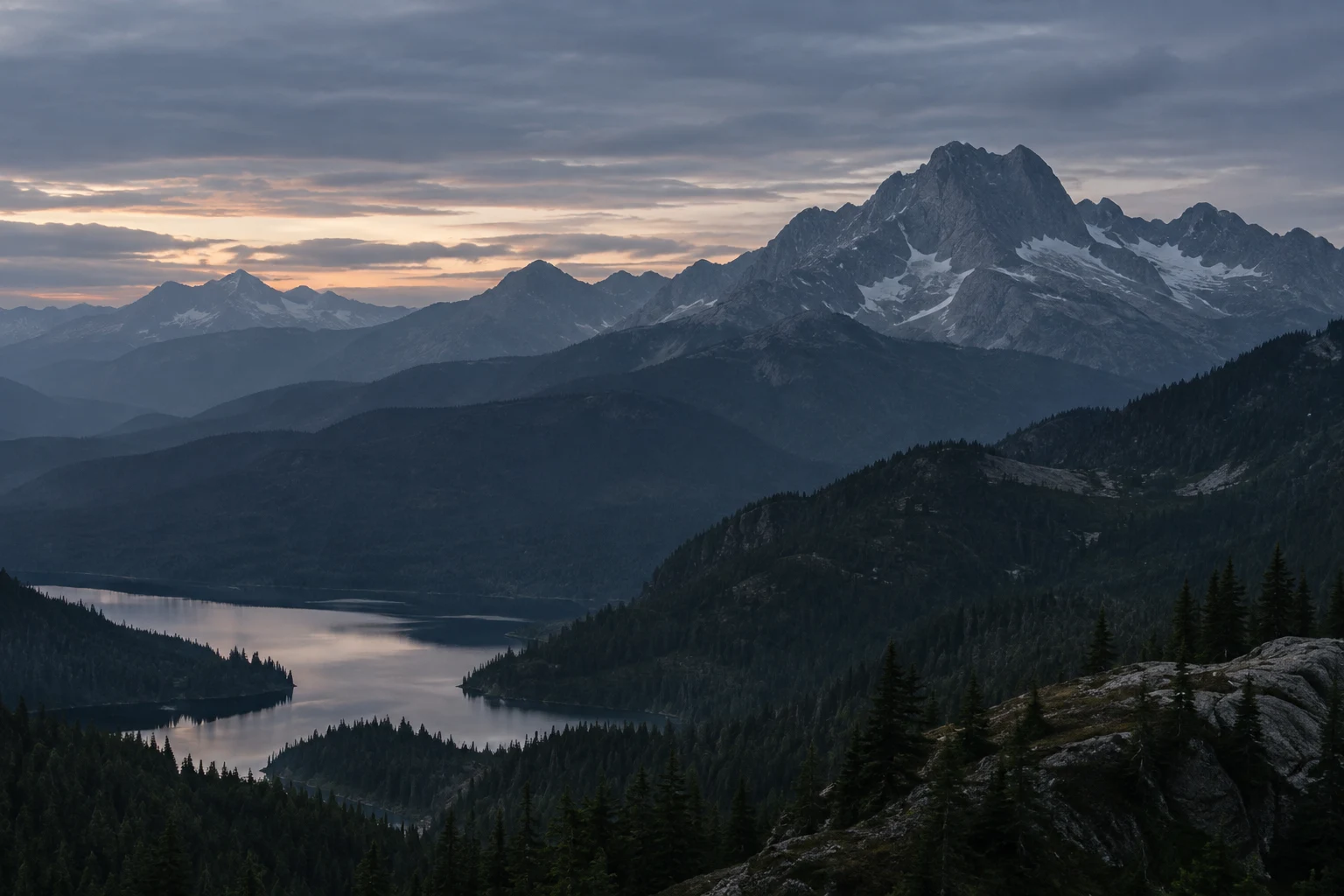 Dark conifer-covered mountain layers surrounding a pale lake beneath a rugged ridge at dusk.