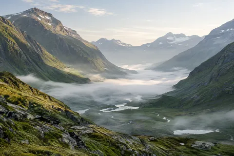 Broad green mountain valley with small water channels and low white mist under pale morning light.