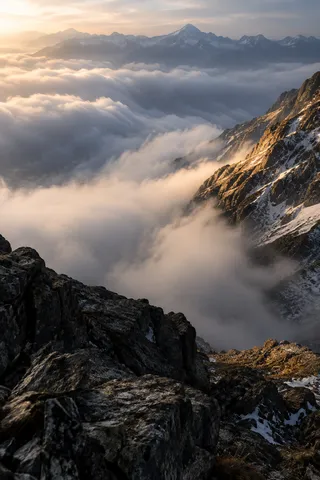 Portrait view down from dark rocks into billowing cloud beside a sunlit mountain wall.