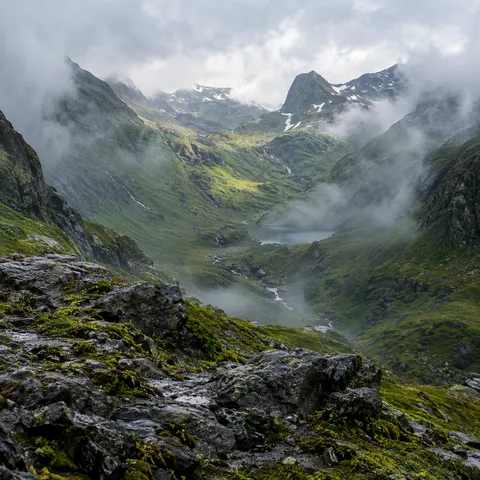 Wet mossy rocks overlooking a green mountain hollow with drifting mist and a small lake.