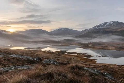 Rounded snowy hills above a misty highland basin with small pools and brown tussock grasses.