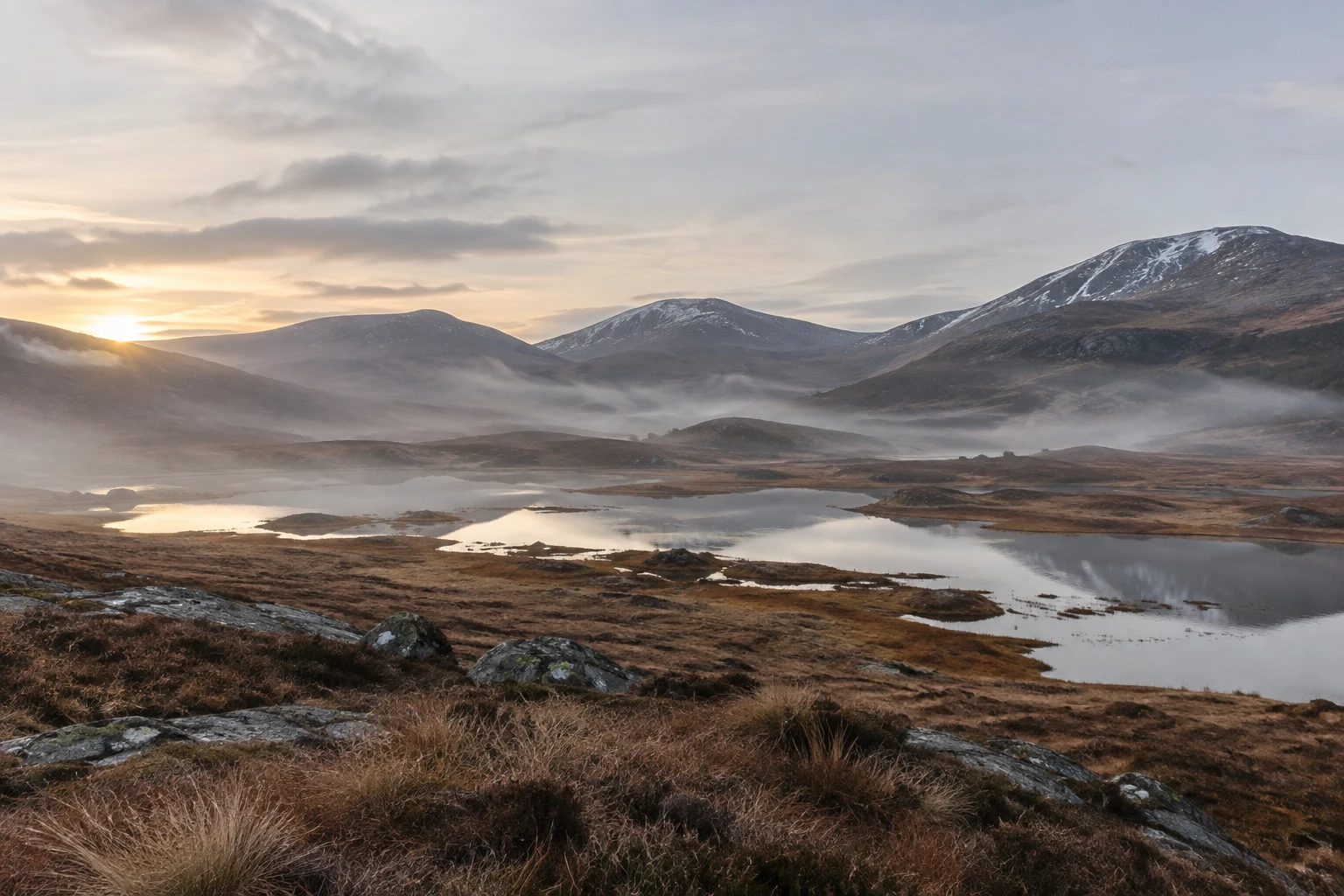 Rounded snowy hills above a misty highland basin with small pools and brown tussock grasses.