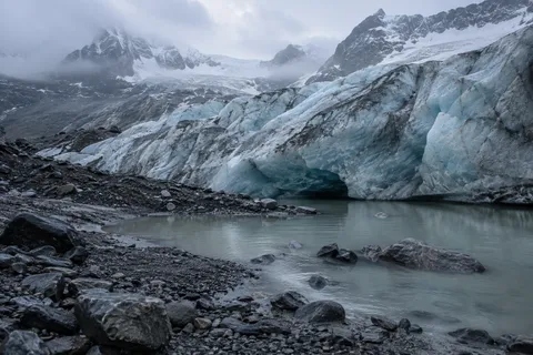 Low glacier edge with blue-white dirty ice, dark gravel and a small gray meltwater pool.