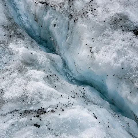 Overhead close view of a curving pale blue groove in rough white glacier ice with dark mineral grains.