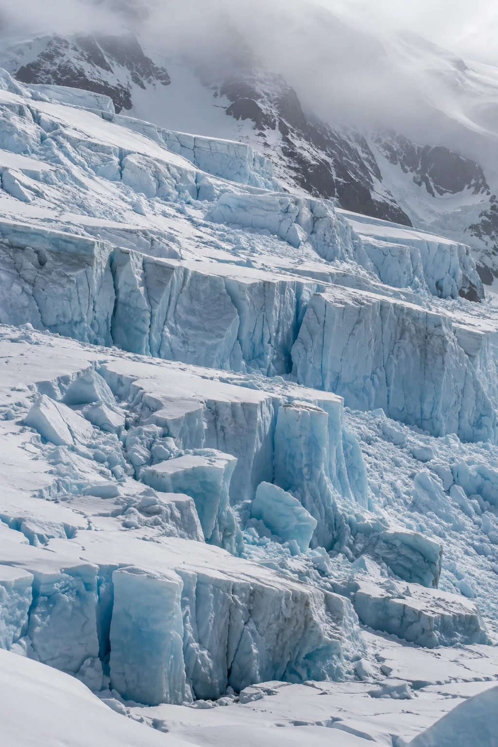 Stepped glacier ice with irregular blue cracks, broken edges and a misty mountain behind.