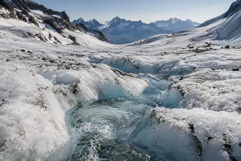 Low view along a clear blue meltwater stream cut into rough white glacier ice.