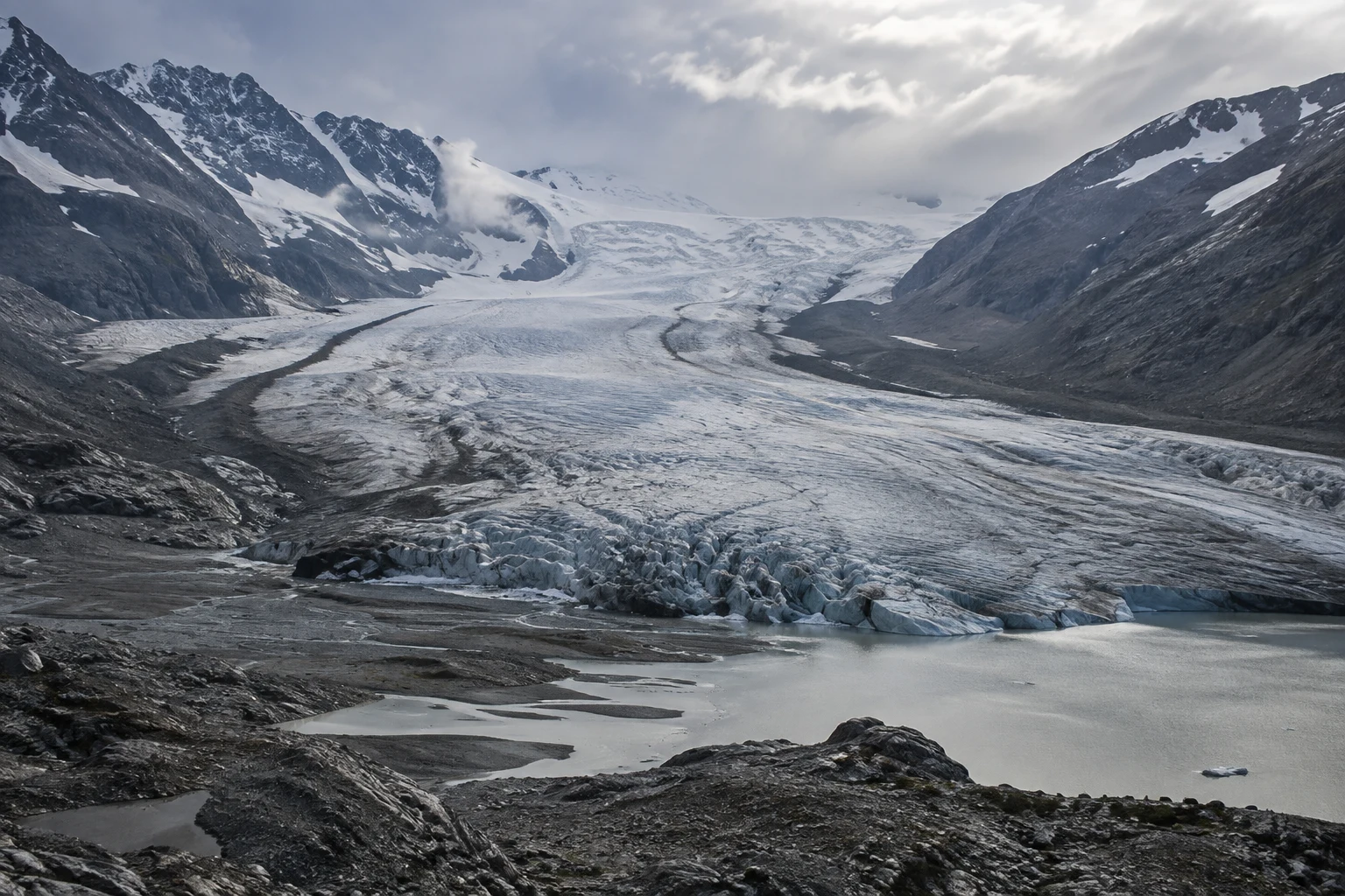 Wide receding glacier tongue between dark mountain slopes with a pale meltwater pool in front.