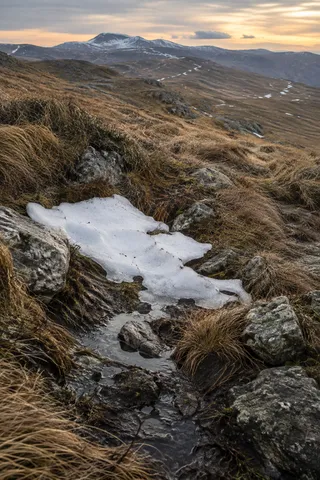 Irregular snow patch between gray stones and brown grass, with a narrow meltwater channel and distant low snowy ridges.