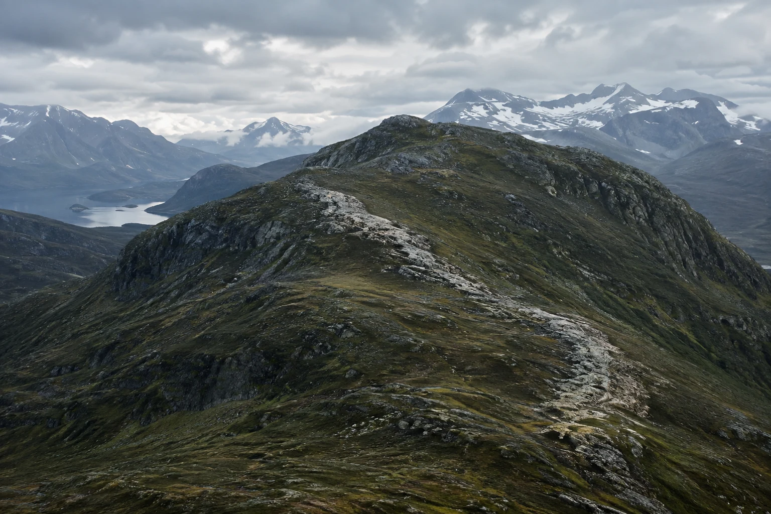Broad rocky foothill with an exposed pale diagonal seam, distant snowy mountains and a small lake at left.