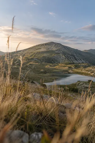 Tall dry grasses and gray stones in soft focus before an olive foothill and small silver-blue lake.