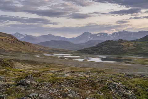 Wide gravel and low scrub basin with connected silver pools between rounded hills and hazy distant mountains.