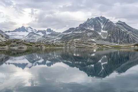 Gray rocky peaks with narrow snow gullies reflected in a calm blue-gray lake beneath an overcast sky.