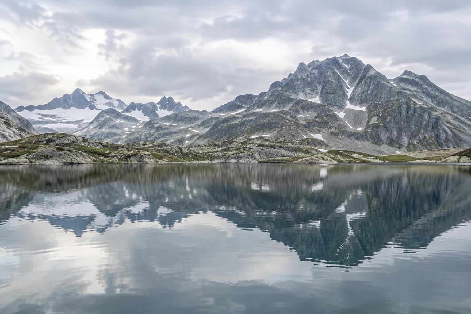 Gray rocky peaks with narrow snow gullies reflected in a calm blue-gray lake beneath an overcast sky.