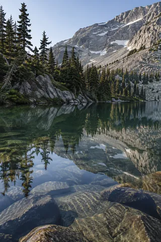 Portrait lake view with pines at left, a pale rocky mountain face above and large submerged stones visible through reflections.