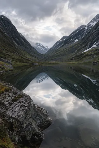 Steep gray-green valley sides and snow patches mirrored in dark water, with a large rock entering at lower left.