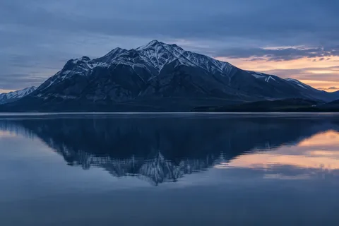 Wide dark blue mountain and its softened lake reflection beneath blue clouds and a small peach opening at right.