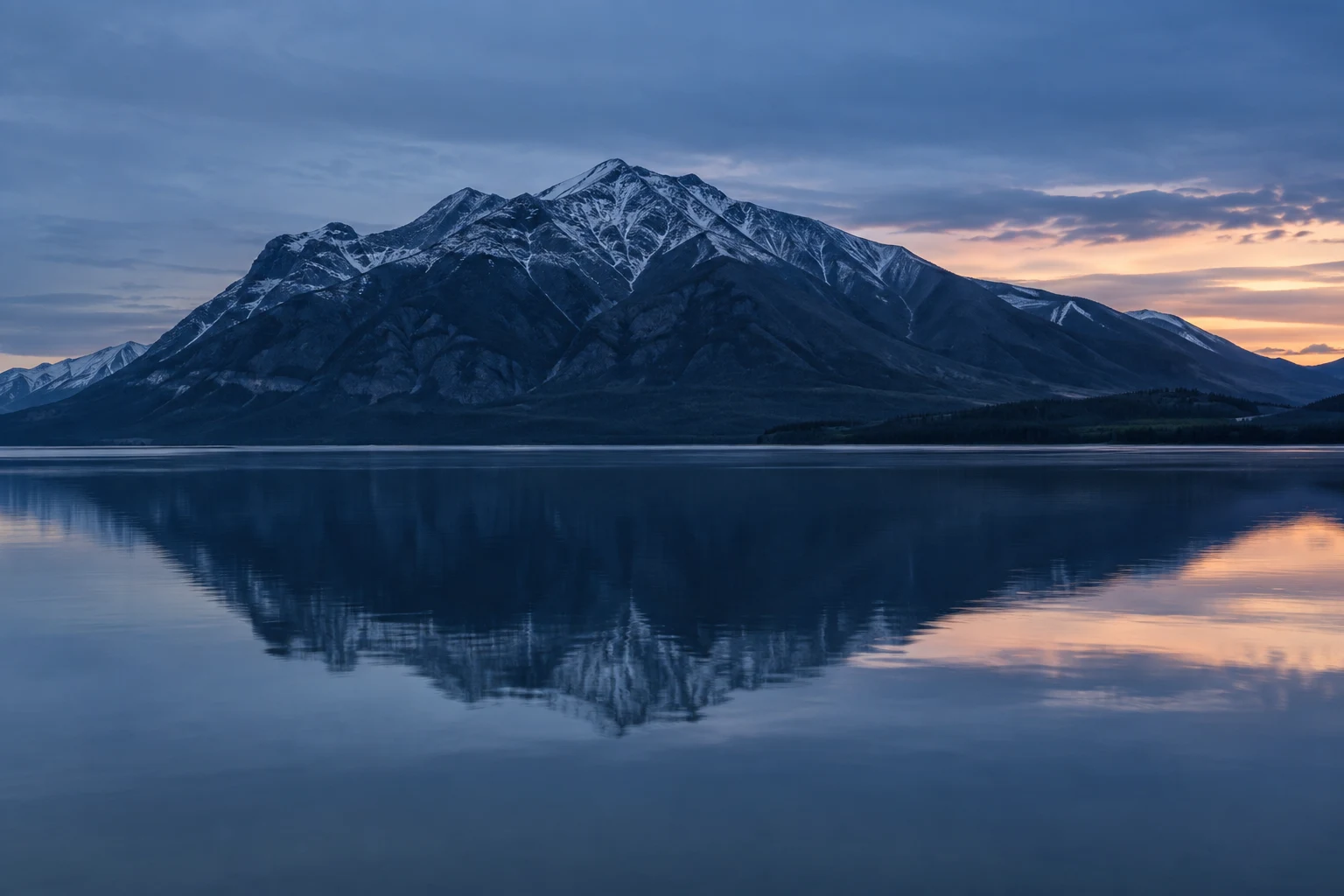 Wide dark blue mountain and its softened lake reflection beneath blue clouds and a small peach opening at right.