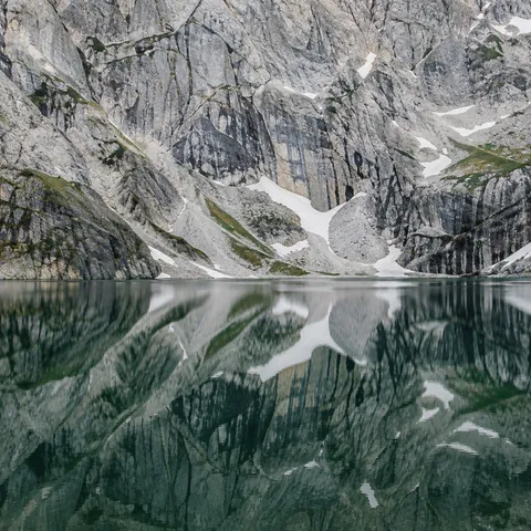 Close view of a fractured gray mountain wall with snow gullies above its green lake reflection, with no sky visible.