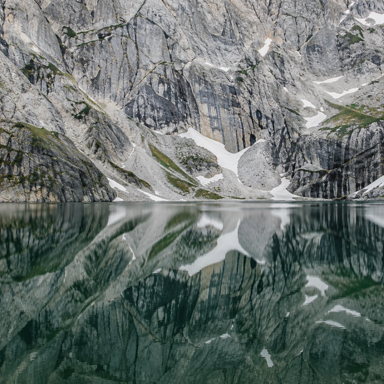 Close view of a fractured gray mountain wall with snow gullies above its green lake reflection, with no sky visible.