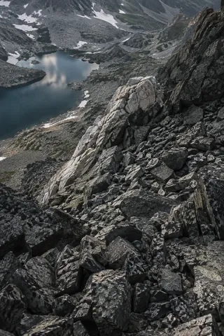 Steep diagonal slope of fractured gray stones with a pale rock spur and a small mountain lake in the upper left.