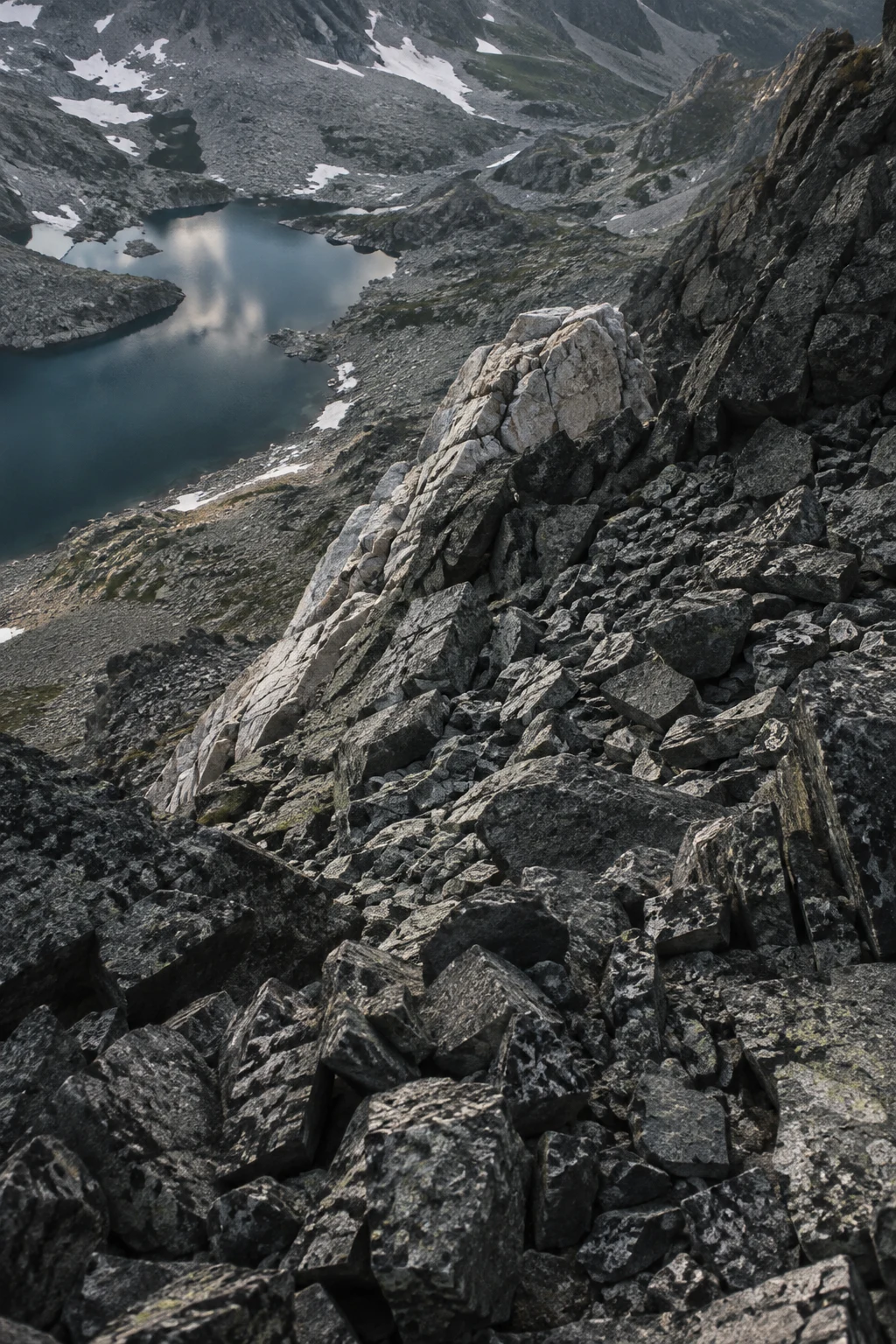 Steep diagonal slope of fractured gray stones with a pale rock spur and a small mountain lake in the upper left.