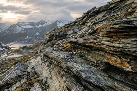 Close diagonal layers of dark slate with traces of ochre lichen and softly focused snowy mountains beyond.